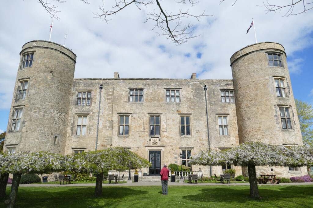 The elevation of a castle with two turrets flanking either side. Several short trees are location in front of the castle.